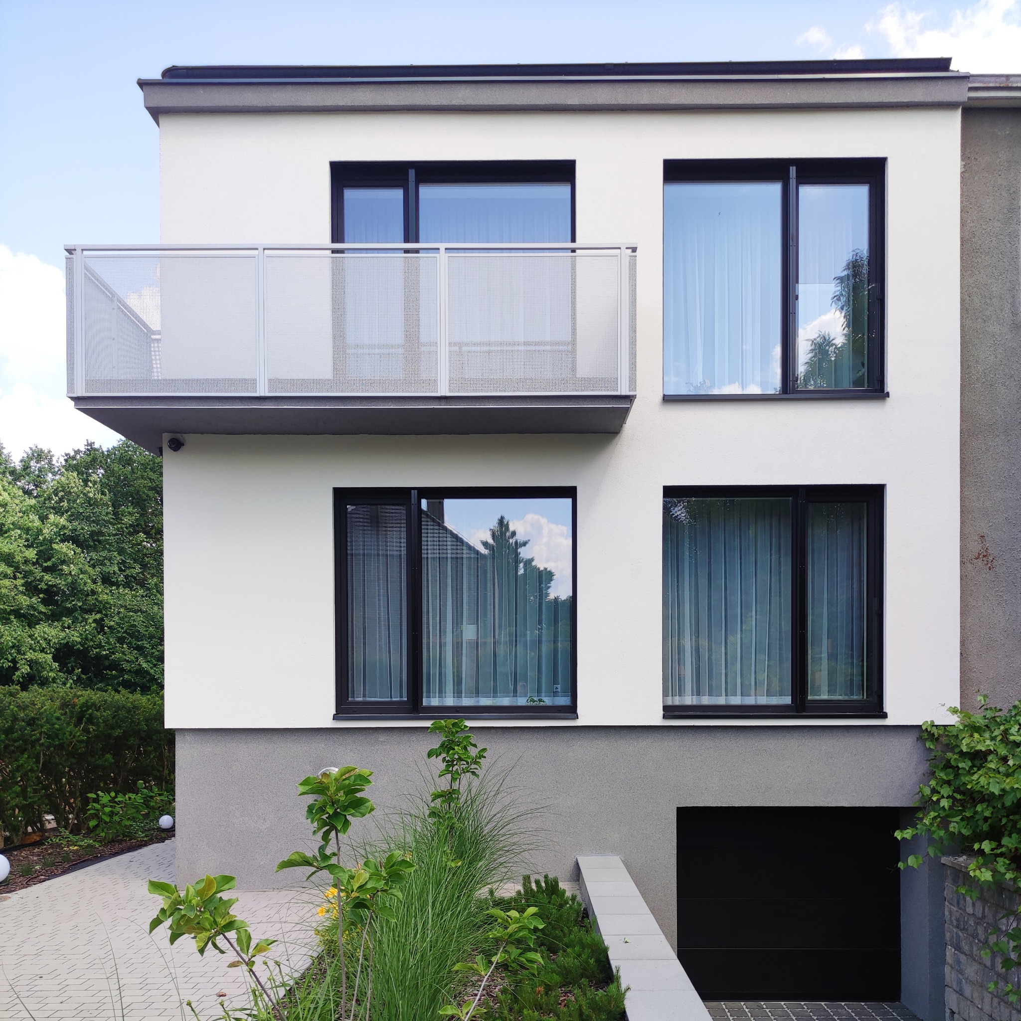 Nowoczesny, dwukondygnacyjny dom z białym tynkiem, czarnymi ramami okiennymi i balkonem z ażurową balustradą. Widoczna brama garażowa, ścieżka z kostki brukowej i roślinność.