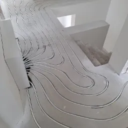 Białe rury instalacji grzewczej ułożone w betonowej wylewce pod ogrzewanie podłogowe, widoczne schody w tle.