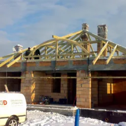 Budowa domu w trakcie zimy, widoczna konstrukcja drewniana dachu z dwoma kominami zabezpieczonymi folią, oraz murowane ściany z pustaków ceramicznych, na pierwszym planie samochód dostawczy z logo...