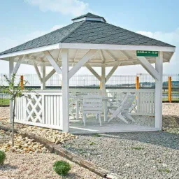 Biała altana ogrodowa z szarym dachem krytym gontem, wyposażona w białe meble ogrodowe, otoczona kamiennym podłożem i młodymi drzewkami.