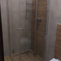 Nowoczesna kabina prysznicowa z transparentnymi ścianami, zamontowana w łazience wykończonej płytkami imitującymi drewno, widok na minimalistyczną armaturę i odpływ liniowy.