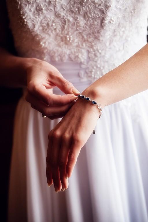 Panna młoda zapina bransoletkę z szafirowymi kamieniami na nadgarstku, na tle białej, zdobionej sukni ślubnej. Delikatne, naturalne światło.