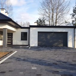 Best Bruk - Nowoczesny dom z ciemną kostką brukową na podjeździe, jasna elewacja i ciemna stolarka okienna, widoczny fragment ogrodu z drzewami bez liści.