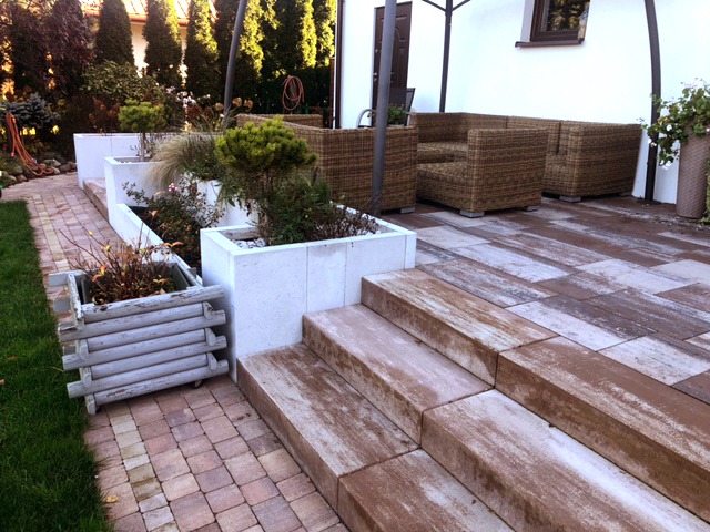 Taras z kostki brukowej imitującej drewno, schody z tego samego materiału prowadzą do strefy relaksu z meblami ogrodowymi z rattanu, w tle rabaty z roślinami w betonowych donicach.