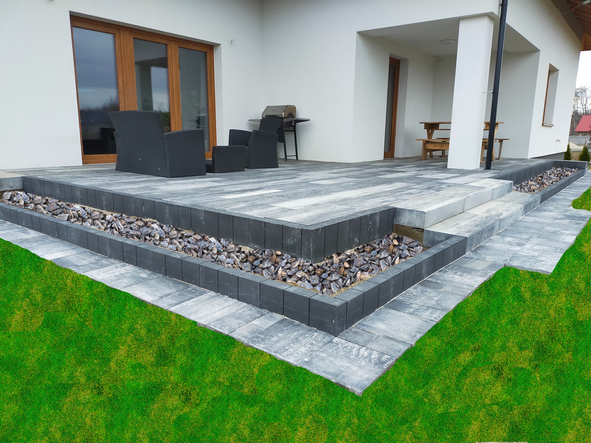 Nowoczesny taras z kostki brukowej w odcieniach szarości, z dekoracyjnym obramowaniem z ciemnych kostek wypełnionych kamieniami, przylegający do białego domu z brązowymi oknami, na tle zadbanego...