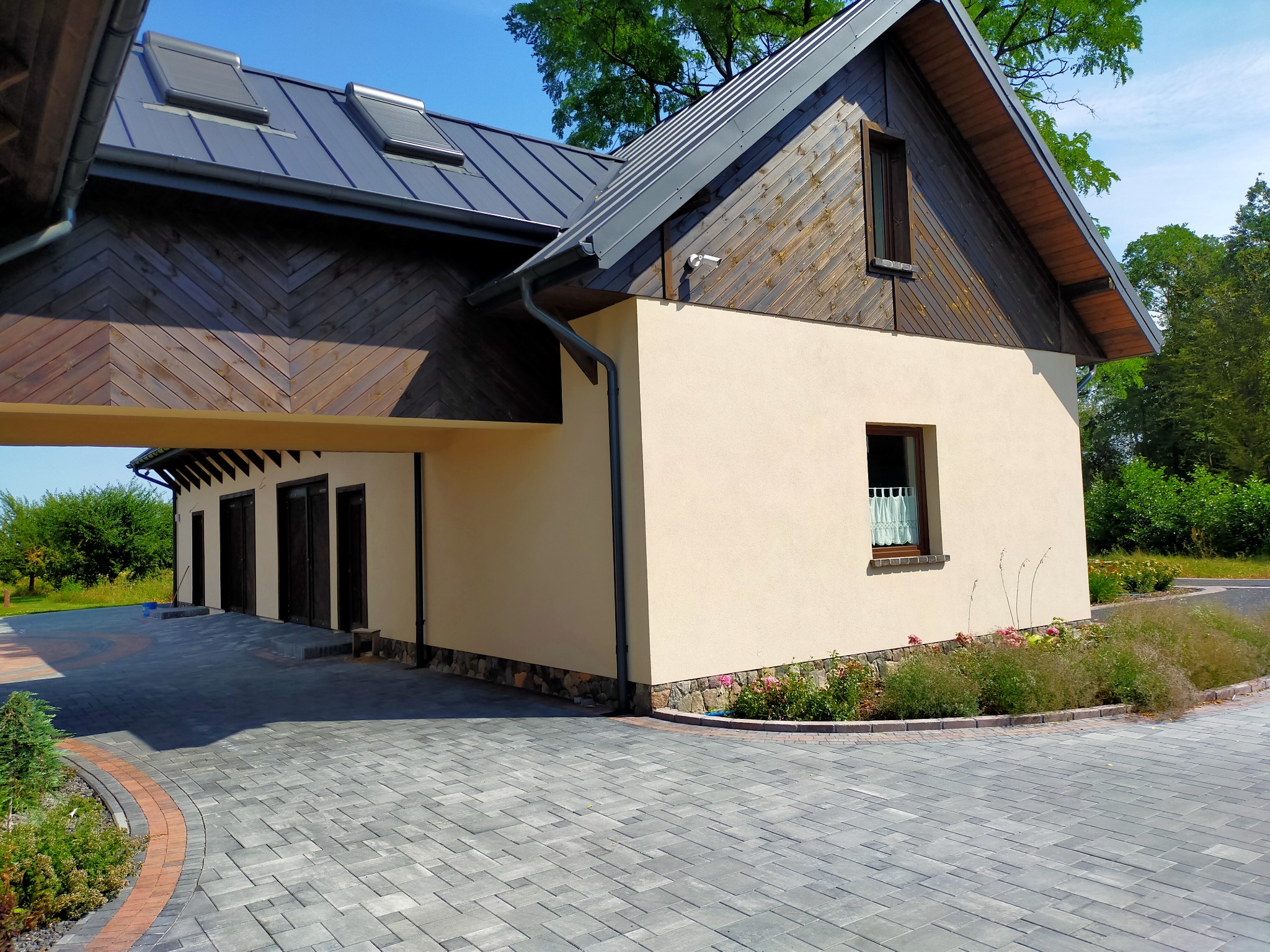 Ułożona szara kostka brukowa prowadząca do garażu z czterema bramami, elewacja budynku w kolorze kremowym z drewnianymi elementami wykończeniowymi, obrzeża z czerwonej kostki.