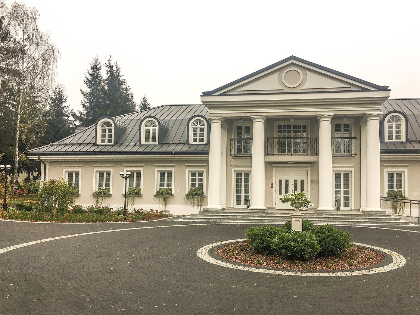 Elegancki dom z kolumnami i brukowanym podjazdem, otoczony zielenią w jesiennej scenerii.