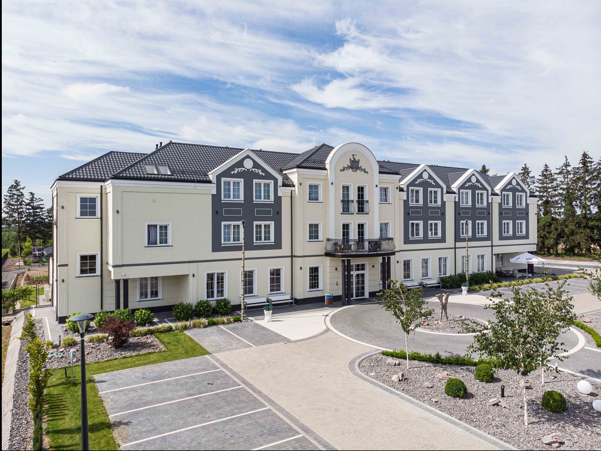 Widok z góry na dwukondygnacyjny budynek z ciemnoszarą dachówką, jasną elewacją i szarymi akcentami, otoczony parkingiem i chodnikiem z kostki brukowej, z elementami zieleni i dekoracyjnymi...