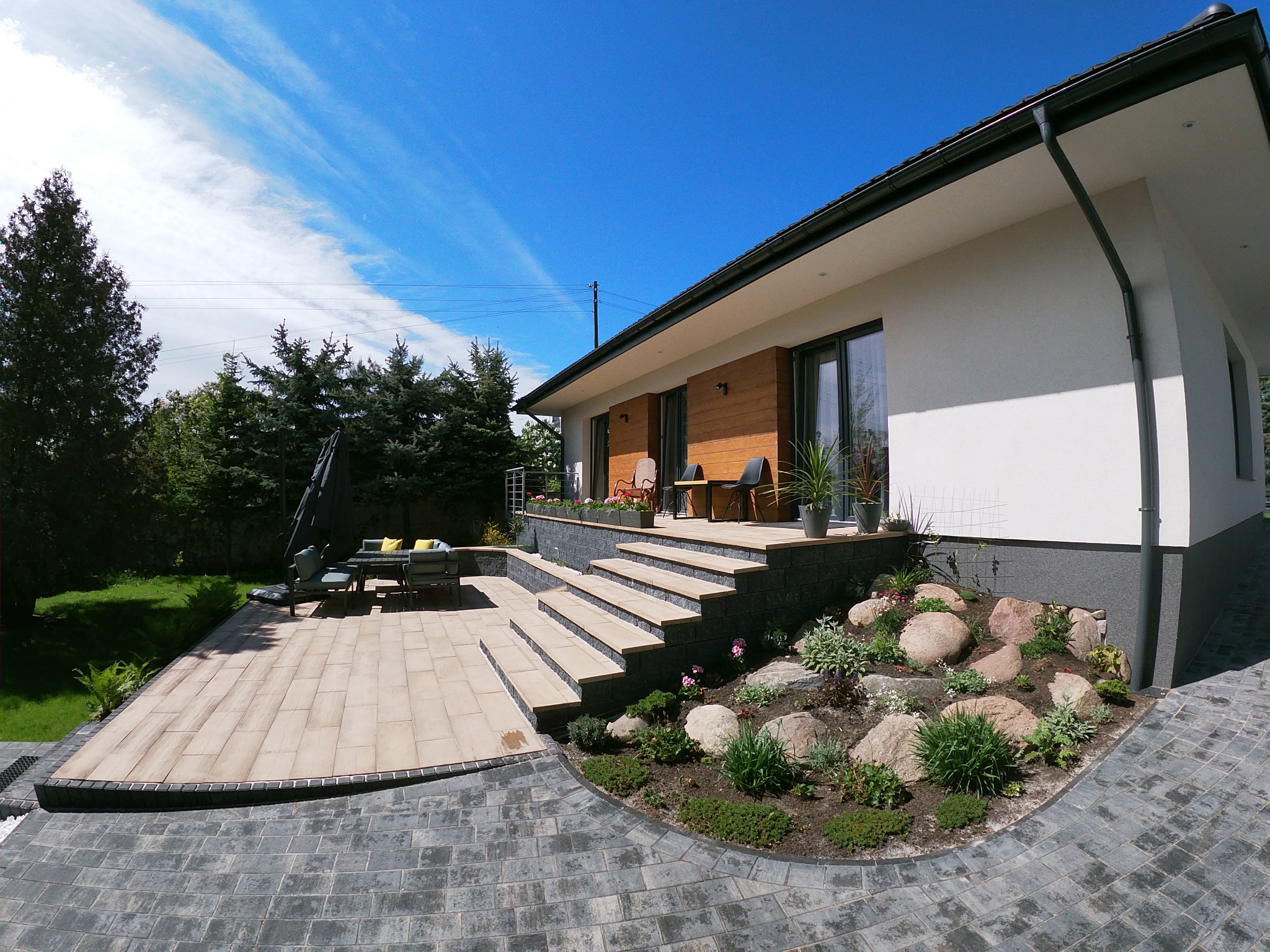 Nowoczesny taras z jasnej kostki brukowej, schody z kamienia prowadzące do domu z drewnianą elewacją, rabata z kamieniami i roślinami, szara kostka brukowa na podjeździe, błękitne niebo.