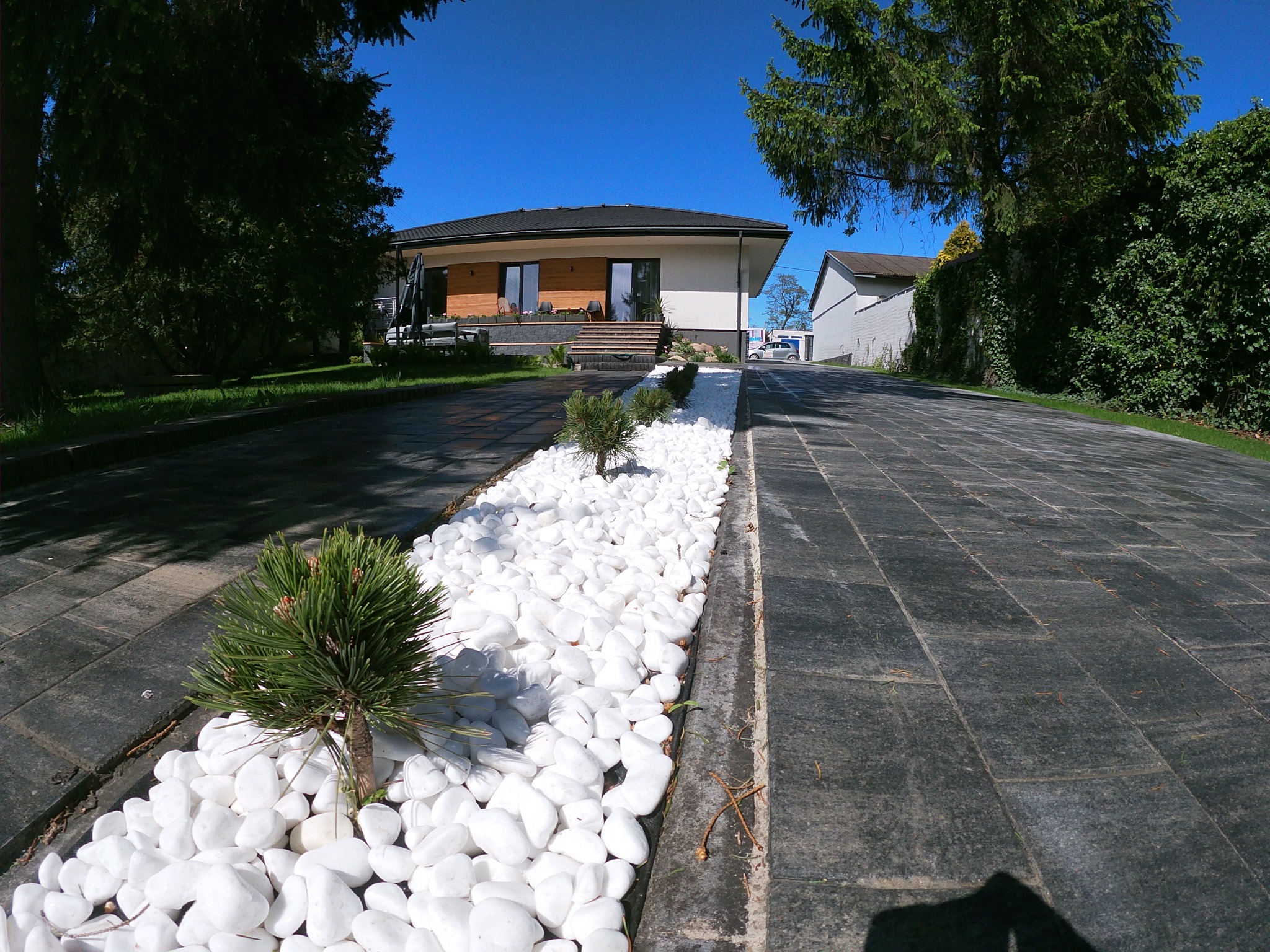 Szeroki kadr na podjazd z ciemnej kostki brukowej, ozdobiony pasem białych kamieni i niskimi iglakami, prowadzący do nowoczesnego domu z drewnianą elewacją w słoneczny dzień.