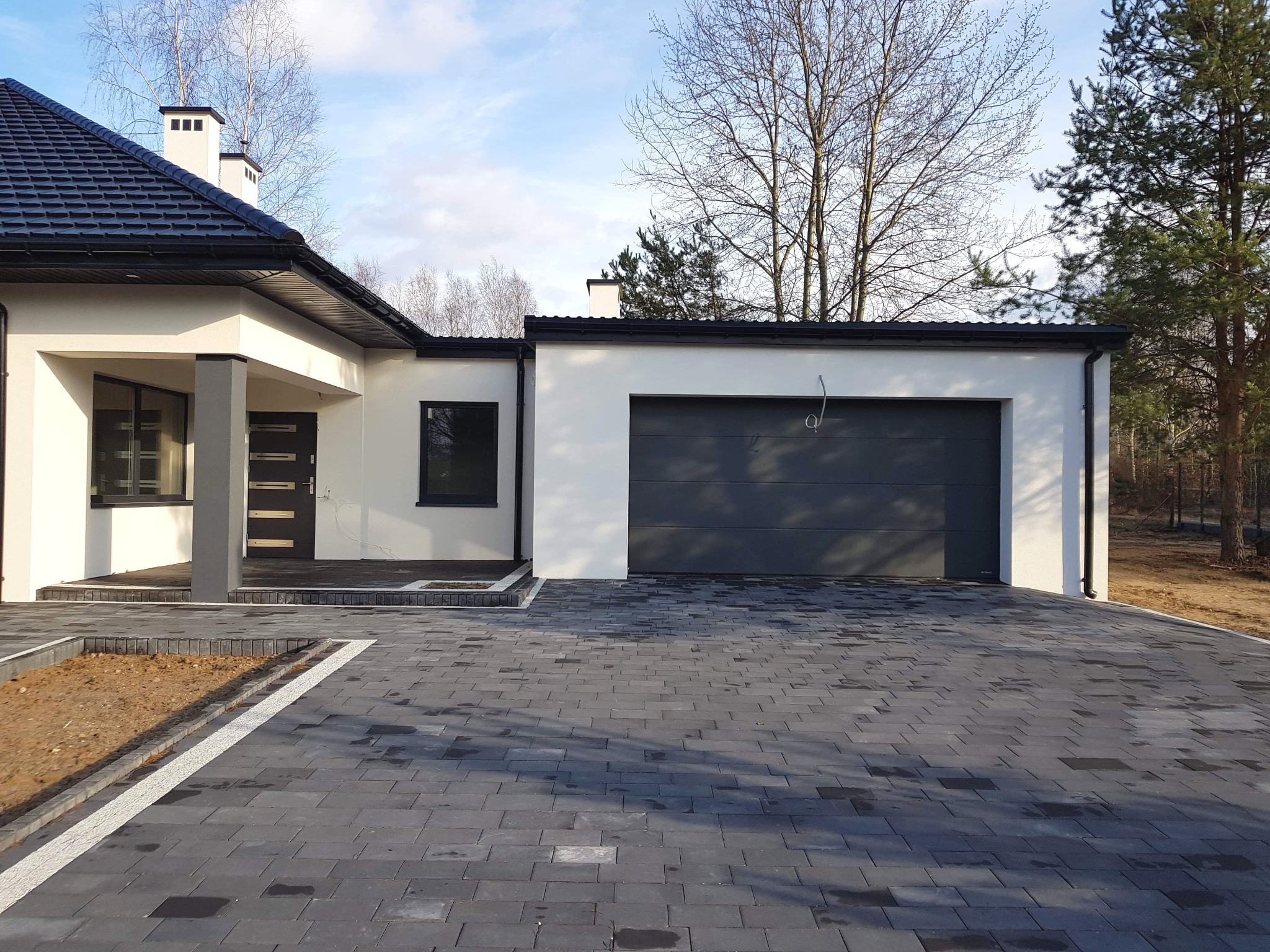 Nowoczesny dom z ciemną kostką brukową na podjeździe, jasna elewacja i ciemna stolarka okienna, widoczny fragment ogrodu z drzewami bez liści.