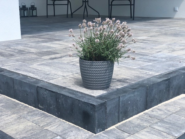Szare kostki brukowe ułożone na tarasie, z doniczką z lawendą w rogu, widoczne czarne obrzeża i meble ogrodowe w tle.