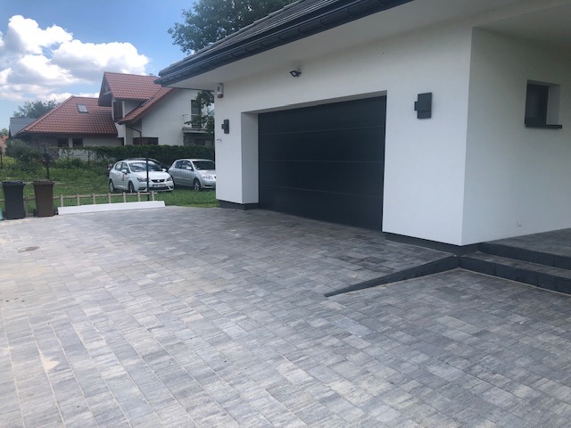 Szara kostka brukowa ułożona przed nowoczesnym domem z czarną bramą garażową i białymi ścianami. Widoczne dwa samochody i domy w tle.