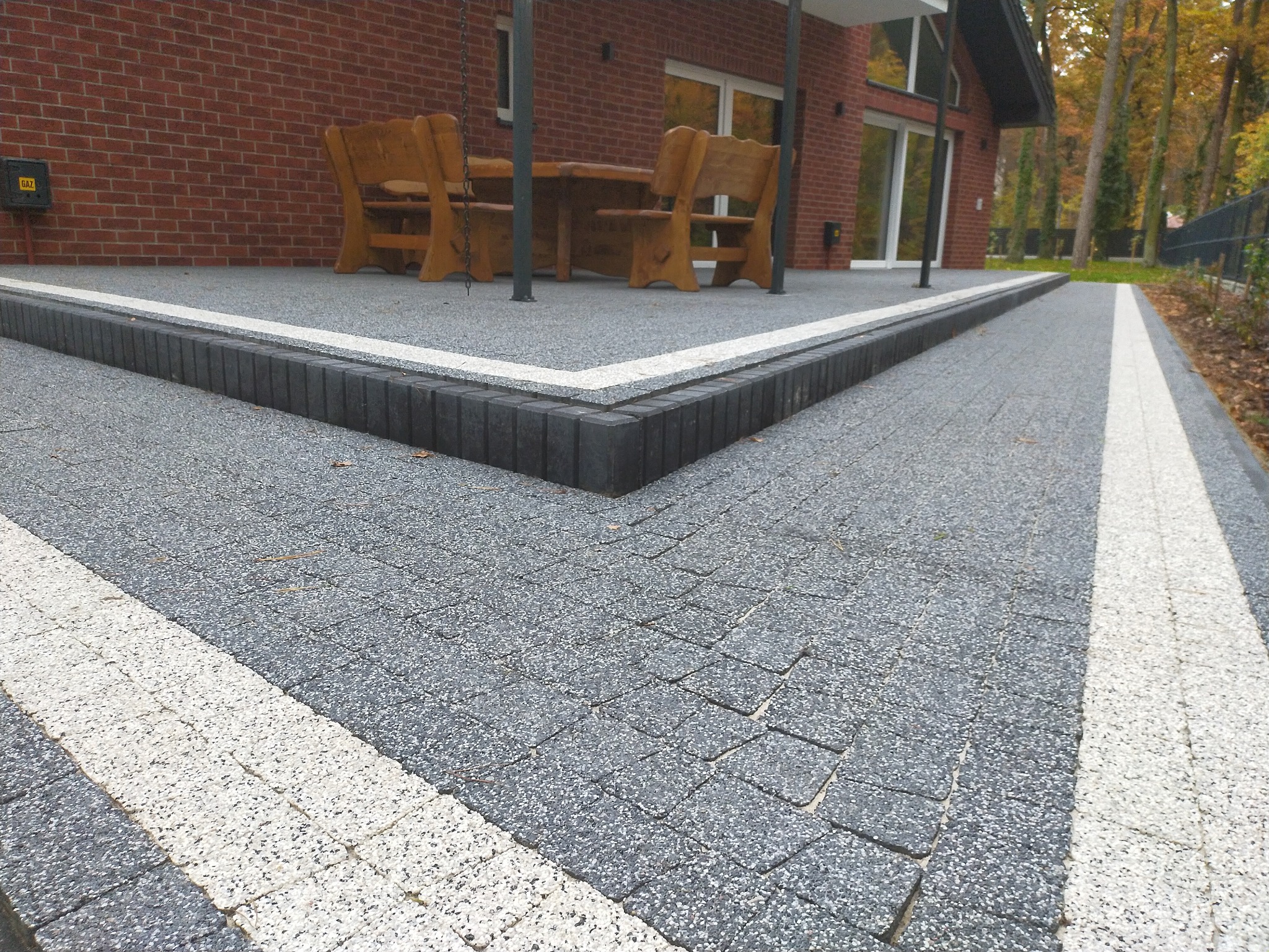 Szara kostka brukowa z białymi pasami oddzielającymi, obrzeża z ciemnej kostki, drewniane meble ogrodowe na tarasie z ceglanym budynkiem w tle.