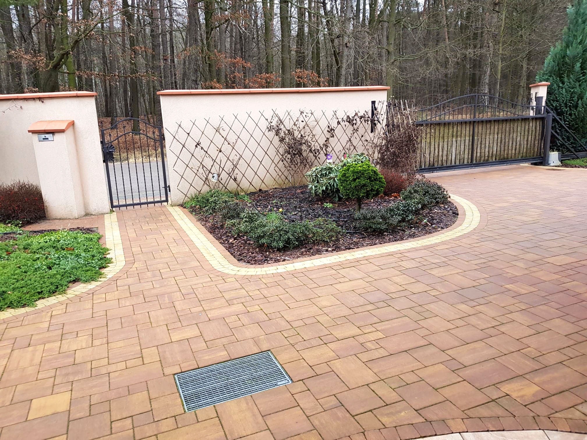 Brązowa kostka brukowa ułożona na posesji z widocznym drenażem liniowym, rabatą z roślinami i ogrodzeniem z bramą w tle.