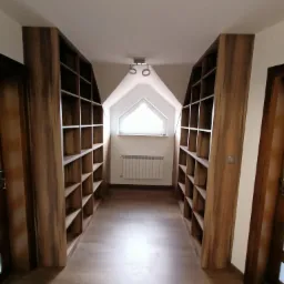 Długi korytarz z symetrycznie umieszczonymi, drewnianymi regałami na książki, dopasowanymi do skosu dachu, z oknem w kształcie trójkąta w tle.