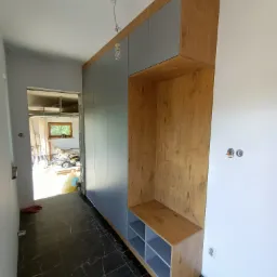 Wysoka szafa w przedpokoju z szarymi frontami i drewnianymi elementami wykończeniowymi, wnęka z półkami i siedziskiem, czarna marmurowa podłoga, widoczne niedokończone wykończenie ścian i sufitu.