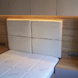 Nowoczesna sypialnia z zabudową: drewniane panele ścienne z oświetleniem LED za wezgłowiem łóżka, tapicerowane wezgłowie z czterech kwadratowych paneli, szafki nocne z gniazdkami elektrycznymi.