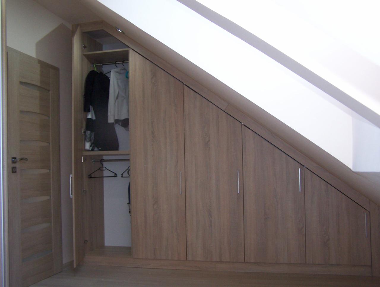 Szafa w zabudowie pod skosem dachu, z frontami w kolorze jasnego drewna, z otwartą częścią z wieszakami i ubraniami.