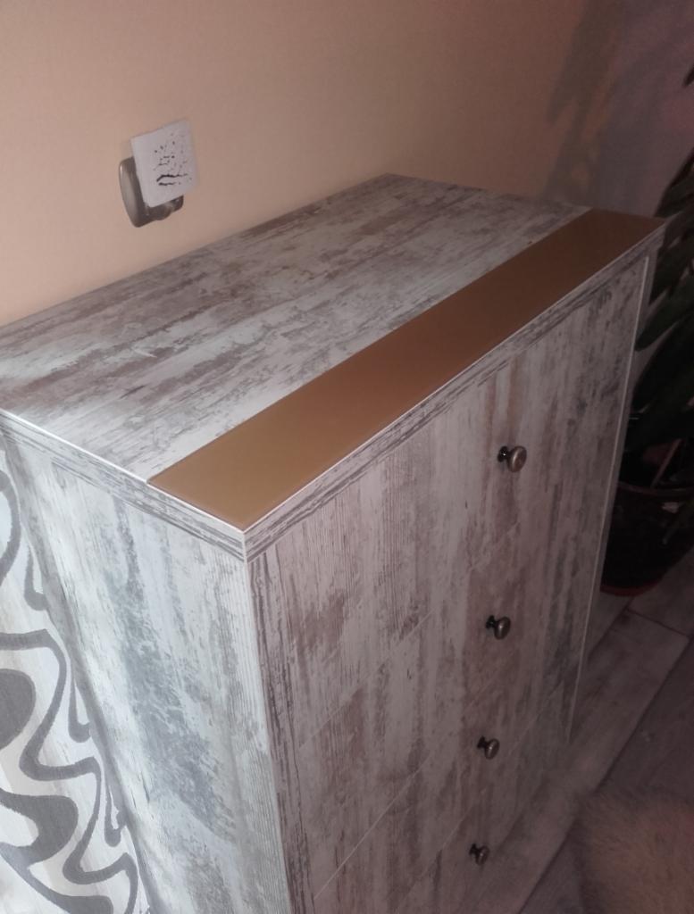 Komoda z szufladami w stylu rustykalnym, z blatem w kolorze miodowego dębu, widoczny fragment ściany i gniazdko elektryczne.