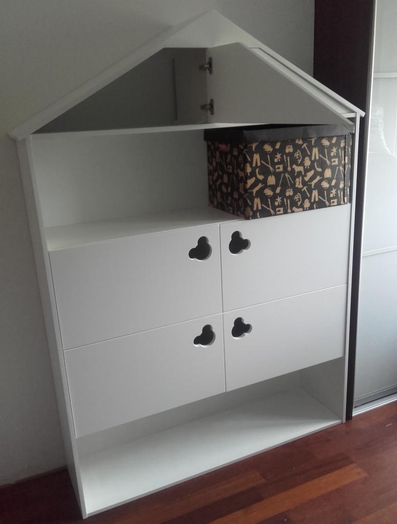 Biała szafka dziecięca na wymiar w kształcie domku z czterema szufladami i pudełkiem na górnej półce, na tle drewnianej podłogi i szafy przesuwnej.