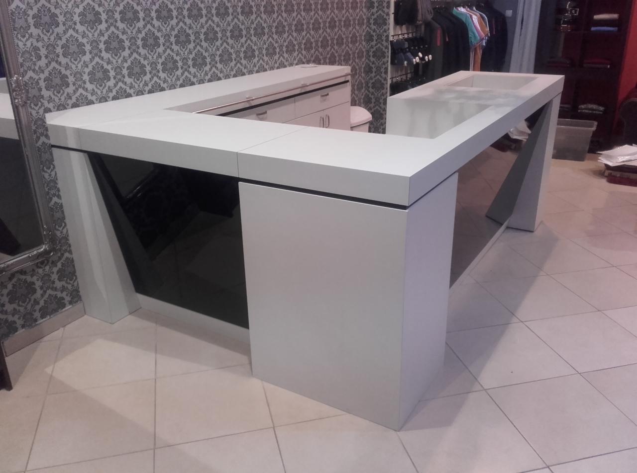 Biała lada recepcyjna w kształcie litery U z czarnymi elementami, widoczna na tle wzorzystej tapety i wieszaków z ubraniami.