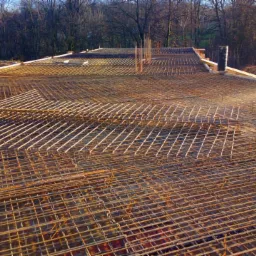 Siatka zbrojeniowa przygotowana do zalania betonem, widoczne pionowe pręty i drewniane szalunki, w tle drzewa i ziemia.