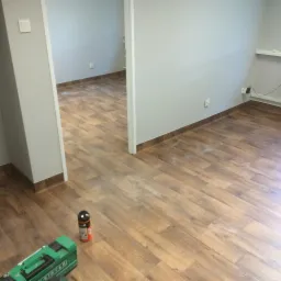 Nowa podłoga imitująca drewno w jasnym pomieszczeniu, widoczna zielona spawarka Leister i klej w tle, listwy przypodłogowe w kolorze ciemnego brązu.