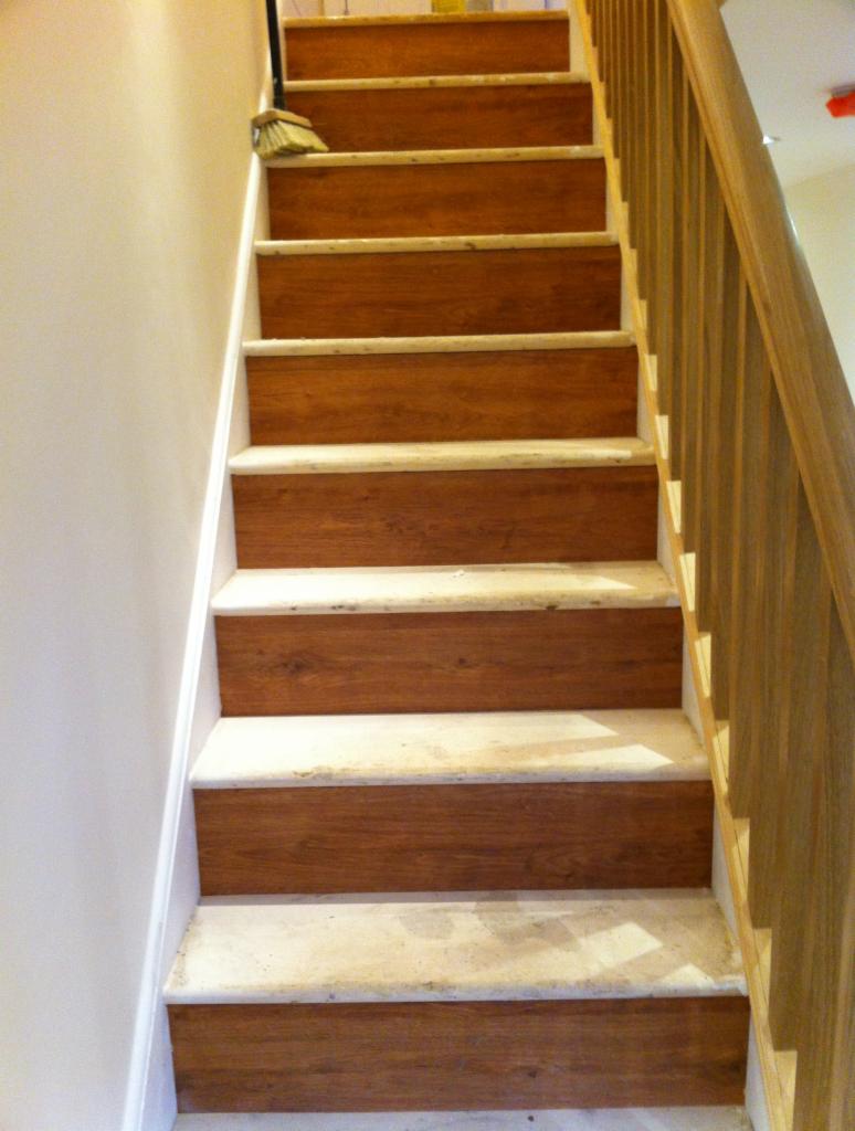 Montaż drewnopodobnych paneli na schodach z białymi podstopnicami i drewnianą balustradą. Na górze schodów stoi miotła. Widok z dołu na górę.