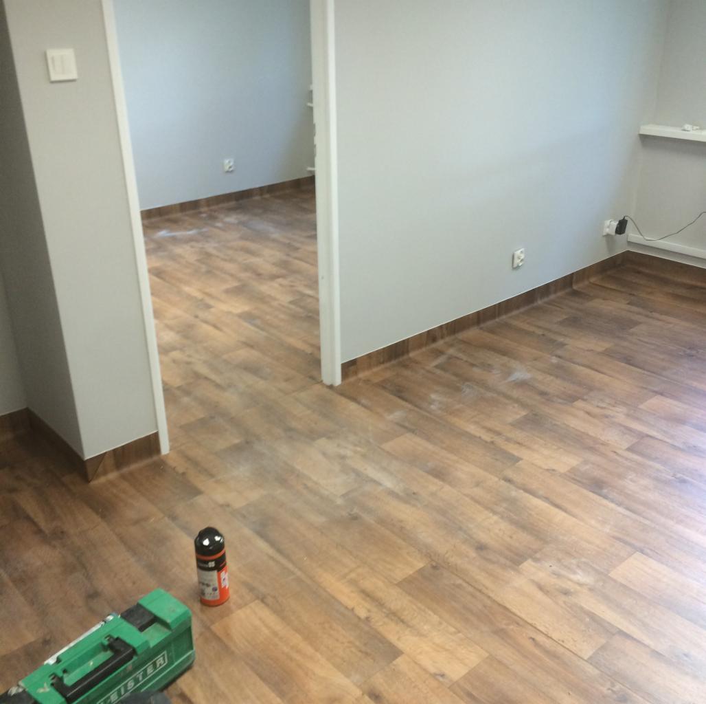 Nowa podłoga imitująca drewno w jasnym pomieszczeniu, widoczna zielona spawarka Leister i klej w tle, listwy przypodłogowe w kolorze ciemnego brązu.