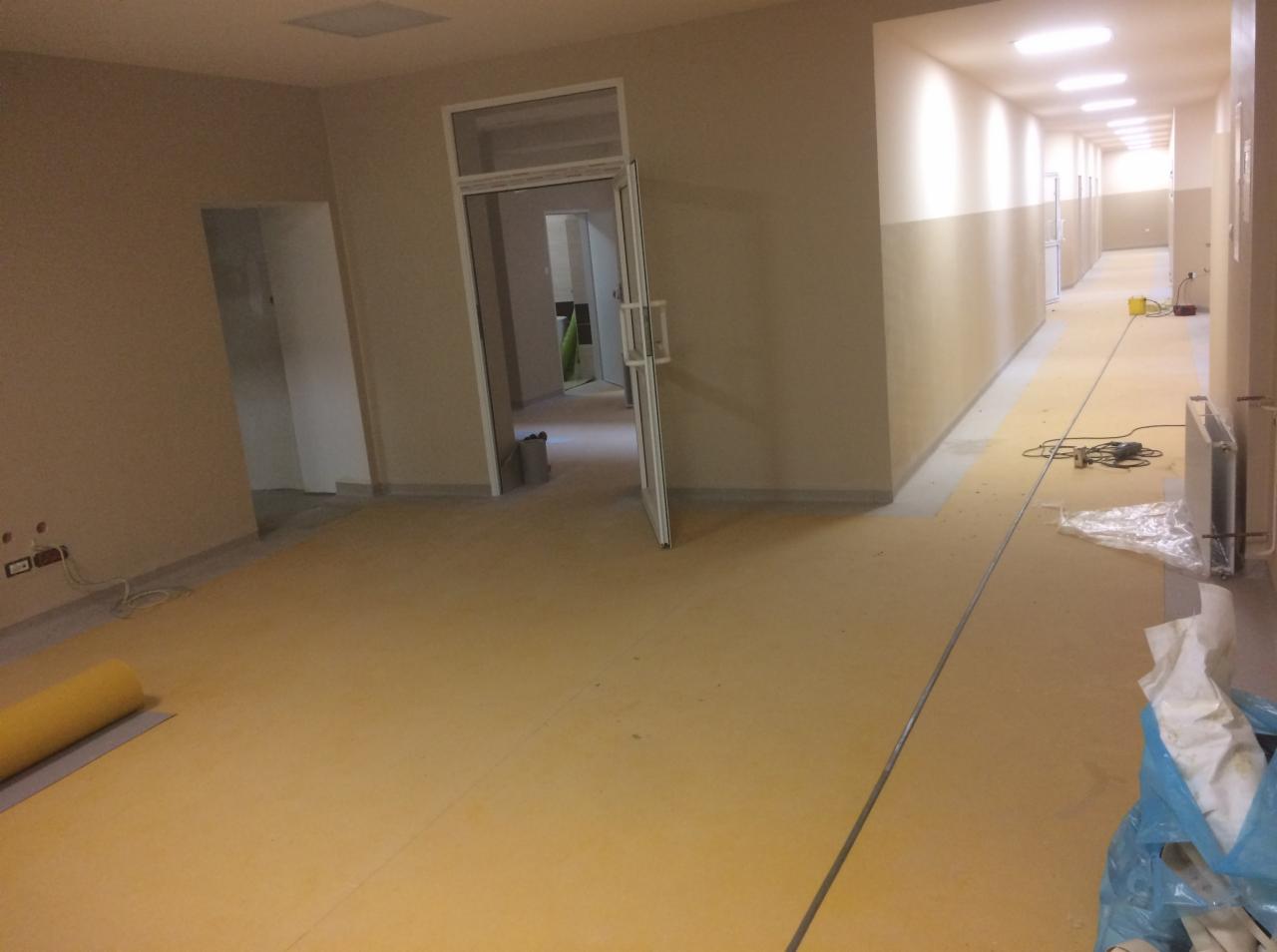 Korytarz w trakcie prac wykończeniowych, widoczna żółta, nowa posadzka oraz narzędzia i materiały budowlane.