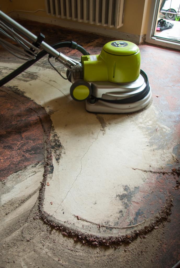 Szlifowanie starej, zniszczonej posadzki maszyną z zieloną obudową, widoczne ślady wcześniejszego wykończenia podłogi.