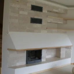 Wykończony kominek z obudową z jasnego kamienia, drewniane półki i kratki wentylacyjne wkomponowane w ścianę, na podłodze duże płytki.