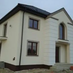 Nowoczesny, dwukondygnacyjny dom z jasną elewacją i ciemnym dachem w Grodzisku Mazowieckim, z widocznymi elementami dekoracyjnymi na fasadzie i brązowymi ramami okiennymi.