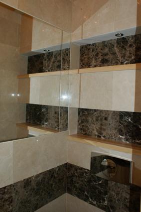 Wykończona łazienka z beżowo-brązowymi płytkami imitującymi marmur, drewnianymi półkami podświetlonymi punktowymi lampami sufitowymi i fragmentem lustra.