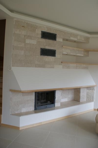 Wykończony kominek z obudową z jasnego kamienia, drewniane półki i kratki wentylacyjne wkomponowane w ścianę, na podłodze duże płytki.