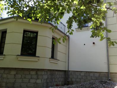 Fragment elewacji domu z widocznym balkonem, oknami z ciemnymi ramami i kamienną podmurówką, częściowo zasłonięty gałęziami drzewa.