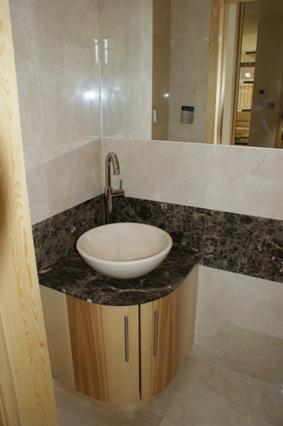 Nowoczesna łazienka z umywalką nablatową w kształcie misy, osadzoną na blacie z ciemnego marmuru i szafką z jasnego drewna o zaokrąglonych krawędziach, wykończona beżowymi płytkami.