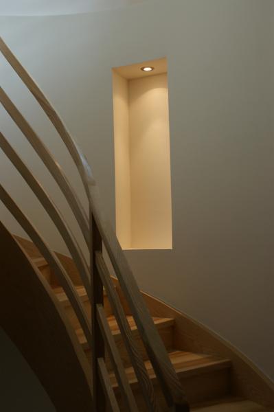 Drewniane schody z zakrętem i balustradą z pionowych listew, z wnęką oświetloną punktowo w ścianie.