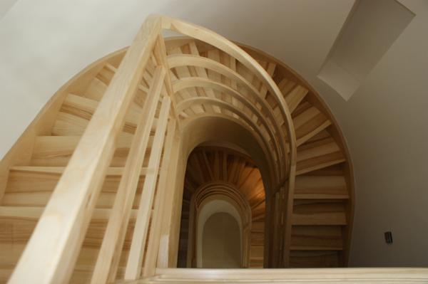 Spiralne schody z jasnego drewna, widok z dołu, ukazujący zakręcony bieg stopni i balustrad w jasnym wnętrzu.