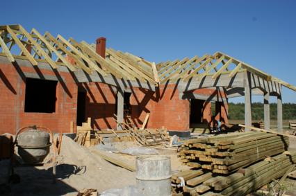 Wznoszenie ścian z czerwonej cegły i konstrukcji drewnianej dachu domu jednorodzinnego w trakcie budowy, widoczne materiały budowlane i betoniarka.