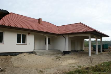 Niedawno wybudowany dom z czerwonym dachem, widoczne fundamenty i teren wokół domu w trakcie prac wykończeniowych, zadaszone miejsce parkingowe po prawej stronie.