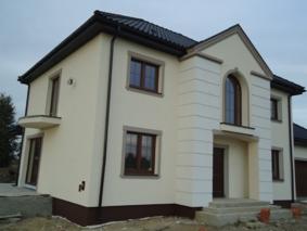 Nowoczesny, dwukondygnacyjny dom z jasną elewacją i ciemnym dachem w Grodzisku Mazowieckim, z widocznymi elementami dekoracyjnymi na fasadzie i brązowymi ramami okiennymi.