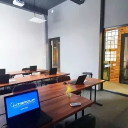 Sala szkoleniowa z drewnianymi stołami, czarnymi krzesłami i laptopami. Na ekranie jednego z laptopów widoczne logo firmy NTGROUP. W tle biała ściana, projektor pod sufitem i tablica suchościeralna.