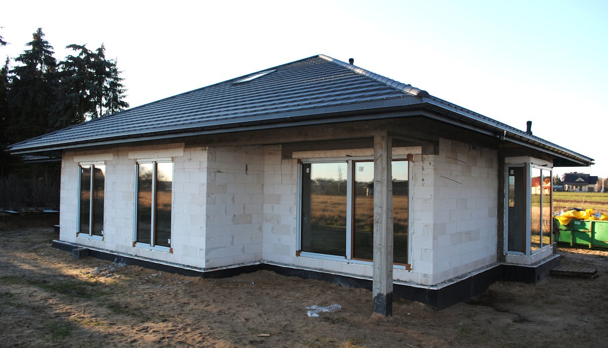 Surowy stan domu jednorodzinnego z białego bloczka, z zamontowanymi oknami z białymi ramami, widoczne odbicie otoczenia w szybach, dach pokryty ciemną dachówką.