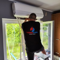 THERMOSTAHL POLAND SP. Z O.O. - Montaż klimatyzacji: mężczyzna na drabinie instaluje biały klimatyzator ścienny przy oknie z widokiem na zieleń. Widoczne narzędzia i logo firmy Thermostahl.