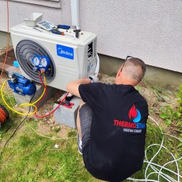 THERMOSTAHL POLAND SP. Z O.O. - Instalacja klimatyzacji Midea: technik w czarnej koszulce z logo firmy Thermostahl podłącza przewody do jednostki zewnętrznej na trawie.