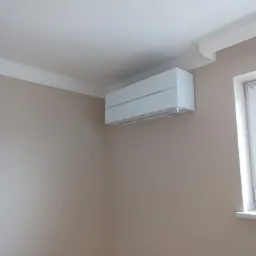 Biały klimatyzator ścienny, model z poziomymi żaluzjami, zamontowany w pokoju z beżowymi ścianami, widoczny fragment okna i dekoracyjny gzyms sufitowy.