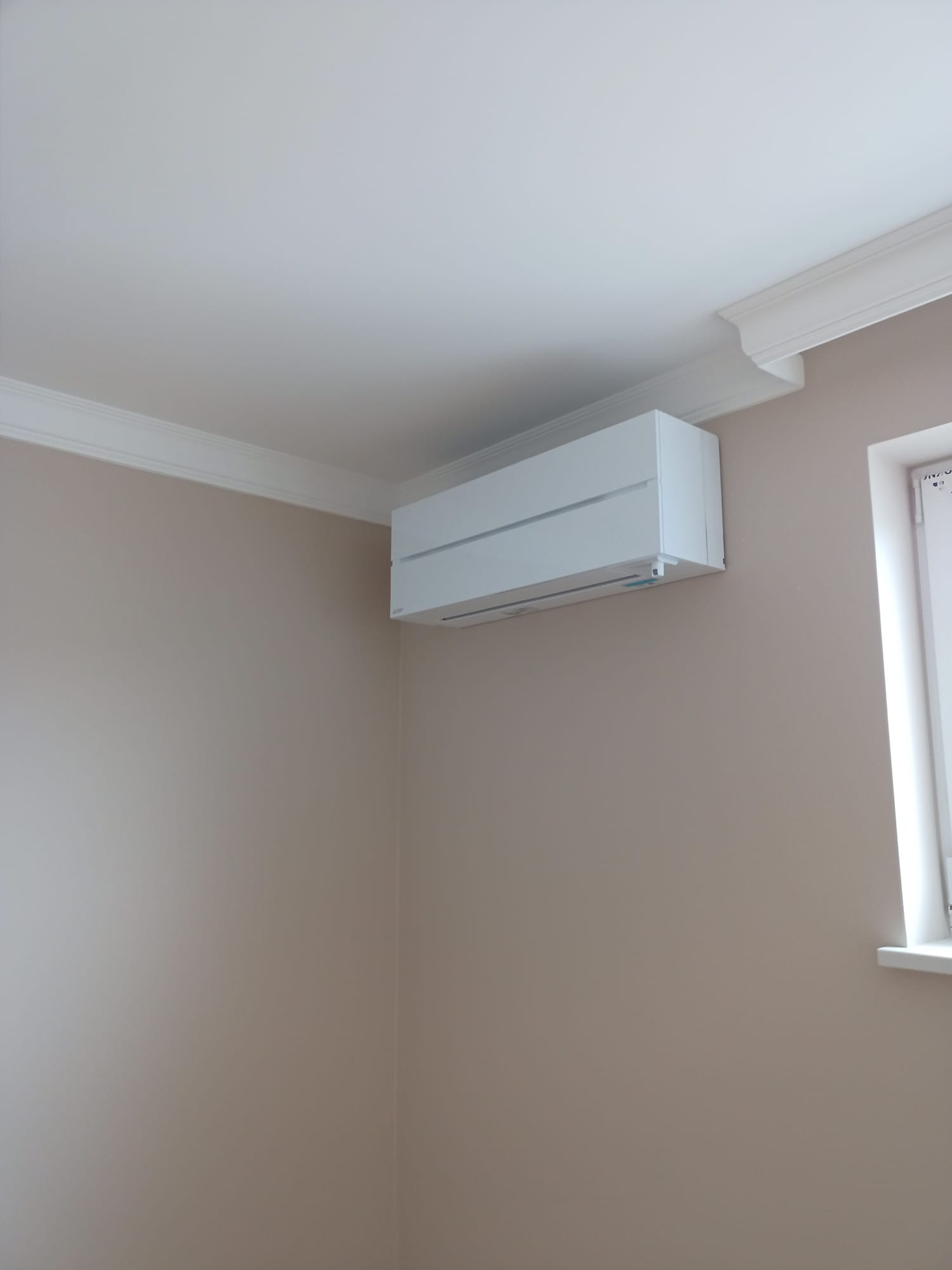 Biały klimatyzator ścienny, model z poziomymi żaluzjami, zamontowany w pokoju z beżowymi ścianami, widoczny fragment okna i dekoracyjny gzyms sufitowy.