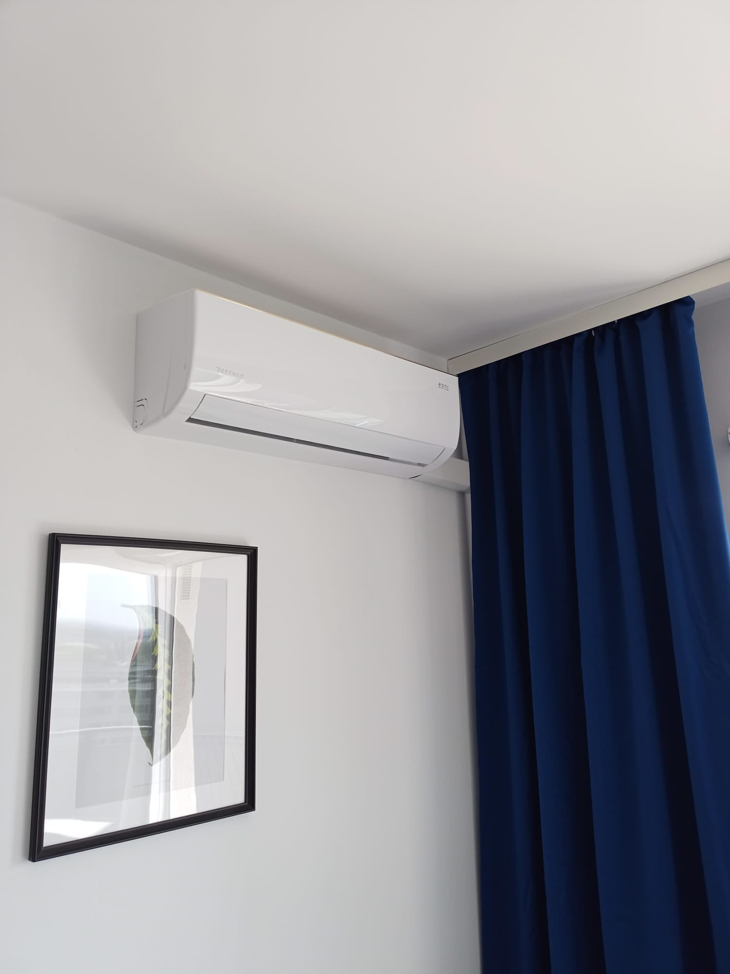Biały klimatyzator ścienny marki Rotenso, zamontowany w jasnym pokoju obok granatowych zasłon i czarnej ramki z obrazem liścia.